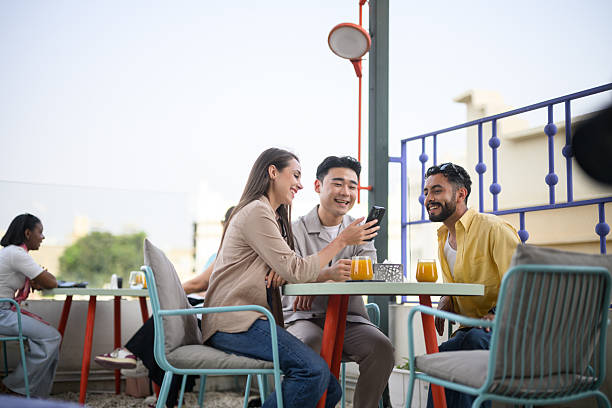 istock Group of young adults using a smartphone together while having drinks at rooftop cafe under clear blue sky 2231505918