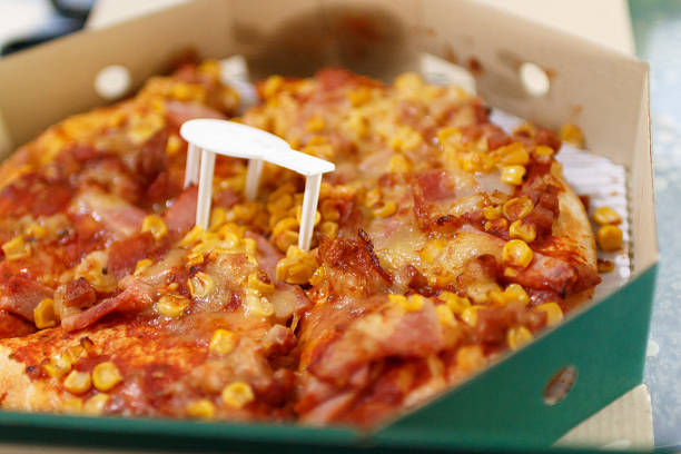Close-up of a hot pizza topped with melted cheese, bacon, and sweet corn inside a cardboard delivery box. A plastic pizza saver is placed in the center to prevent the lid from touching the toppings. Perfect for illustrating concepts of fast food, takeout, delivery service, comfort food, and casual dining.