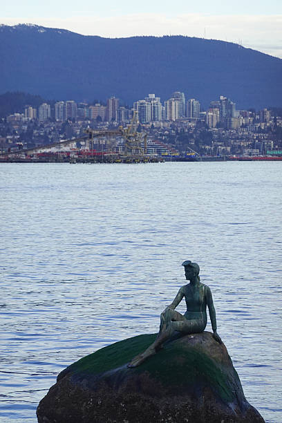 The bronze Girl in a Wetsuit sits on a seaweed stained rock near the seawall, a playful echo of a mermaid in practical gear. Gentle waves and distant skyline forms place the figure within Vancouver’s working harbor. Texture on bronze and stone remains clean, making the photo useful for cultural features and visitor information.