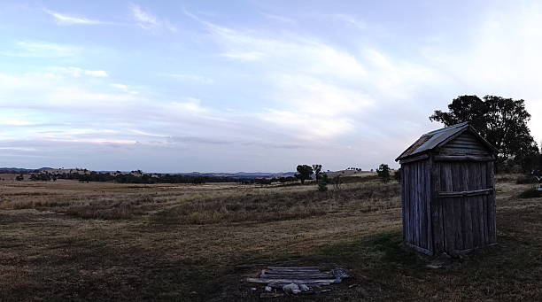 A weathered and ramshackle outhouse stands alone in an arid rural paddock, surrounded by dry grasses and scrub under a cloudless sky in the harsh Australian outback.