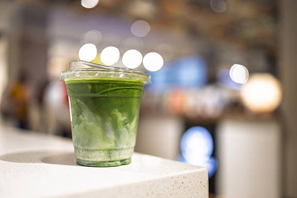 Close-up of iced matcha green tea latte in a clear plastic cup with lid, placed on a counter indoors.
