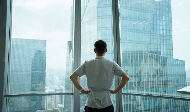 istock Back shot: Man looking out the window at a city office building 2231587490