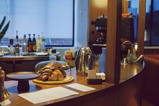 Close-up of fresh croissants under a glass dome on a retro-style coffee bar counter, with coffee machine, drinks, and menu in the background. Location: café interior.