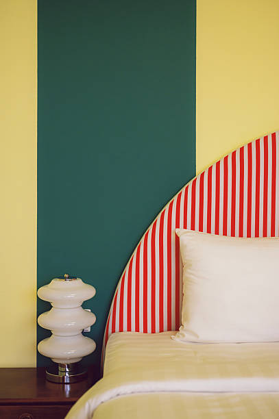 Interior design detail of a retro-style bedroom featuring a red striped headboard, white bedding, bedside lamp, and bold green and yellow color-block wall.