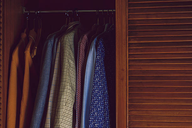 Row of shirts and jackets hanging on hangers inside a partially open wooden wardrobe, showing organized clothing storage in a retro interior.