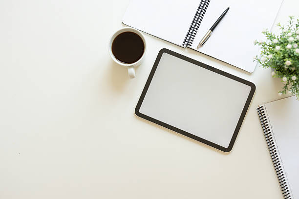 Table top view of digital tablet, notebook, eyeglasses and coffee cup on white table