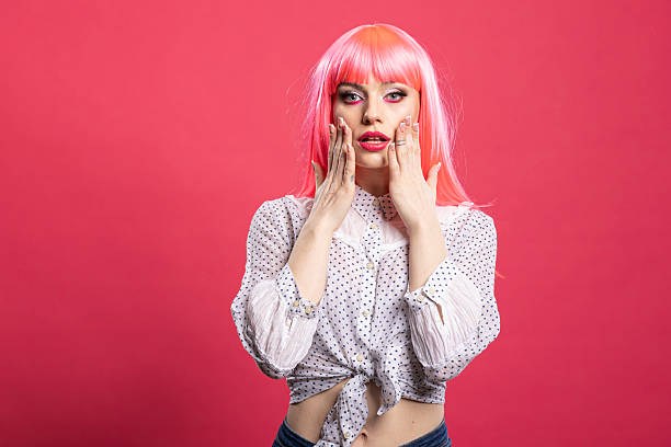 Attractive caucasian woman touching her cheeks and looking at camera with shocked facial expression. Surprised female individual standing with wide eyes and hands on face against pink background.
