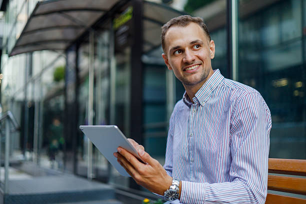 istock Young businessman sitting on a bench, smiling while using a digital tablet, enjoying the outdoors in a vibrant urban setting 2231614117