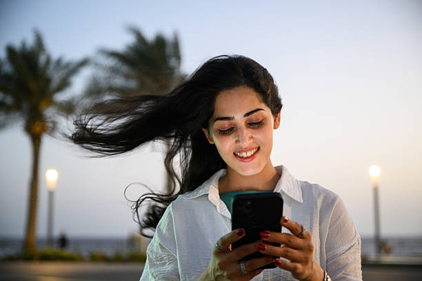istock Smiling young woman with windblown hair using smartphone on Jeddah Corniche promenade during breezy sunset 2231614120