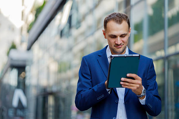 istock Businessman wearing blue suit using digital tablet while walking outside modern office building 2231615572