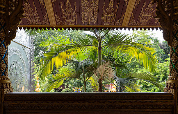 A wide shot of a traditional Buddhist temple's intricate roof, partially hidden by lush green foliage in the foreground. The roof is made of red tiles and features stunning golden decorations with detailed carvings of elephants and other figures. The temple is set amidst a tropical environment with dense green trees and palm fronds surrounding it. The cloudy sky provides a soft, even light that highlights the architectural beauty and vibrant colors of the structure, conveying a sense of peace and natural harmony.