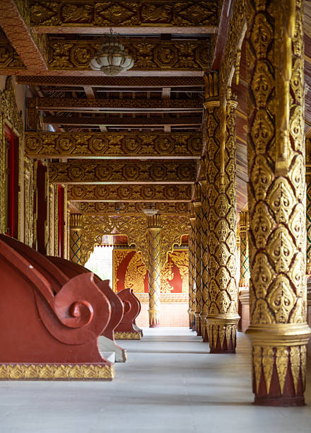 A detailed close-up shot showcasing the intricate and ornate architecture of a Buddhist temple. The foreground is filled with stunning golden carvings and motifs on a reddish-orange wall. The focus is on a square window-like opening that reveals a glimpse of the temple's interior, where a large, cylindrical red pillar with golden patterns stands on a golden base. The vibrant colors and meticulous craftsmanship highlight the rich cultural and spiritual heritage of the region. The natural light accentuates the depth and texture of the carvings, creating a captivating and beautiful image.