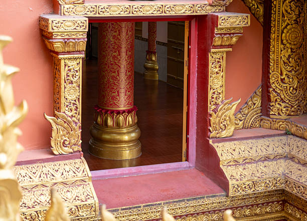 A detailed close-up shot showcasing the intricate and ornate architecture of a Buddhist temple. The foreground is filled with stunning golden carvings and motifs on a reddish-orange wall. The focus is on a square window-like opening that reveals a glimpse of the temple's interior, where a large, cylindrical red pillar with golden patterns stands on a golden base. The vibrant colors and meticulous craftsmanship highlight the rich cultural and spiritual heritage of the region. The natural light accentuates the depth and texture of the carvings, creating a captivating and beautiful image.