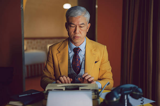 senior chinese man in a suit typing on a vintage typewriter at a desk with an old rotary phone beside him, lit by soft window light for a classic, cinematic office mood. Location: indoor retro hotel suite room
