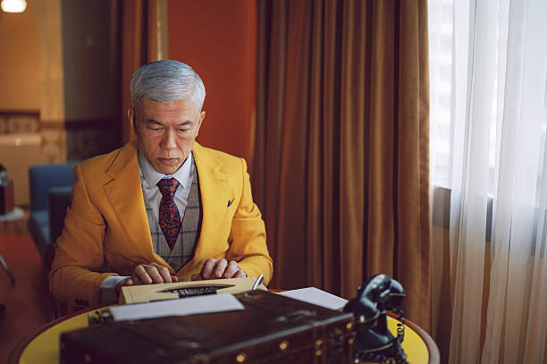 senior chinese man in a suit typing on a vintage typewriter at a desk with an old rotary phone beside him, lit by soft window light for a classic, cinematic office mood. Location: indoor retro hotel suite room