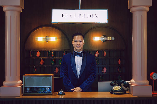 Asian professional male hotel receptionist in a bow tie and velvet suit standing confidently at the front desk under a glowing reception sign. Retro-inspired interior design with key wall, vintage telephone, and classic décor elements. Concept of hospitality, concierge service, and luxury hotel welcome. Location: hotel lobby, Asia.