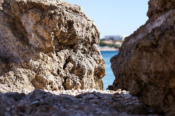 Close-up of textured coastal rocks forming a natural frame that opens up to a serene sea view in the distance. The sharp light, earthy tones and hint of beach beyond create a balanced composition full of summer and exploration mood.