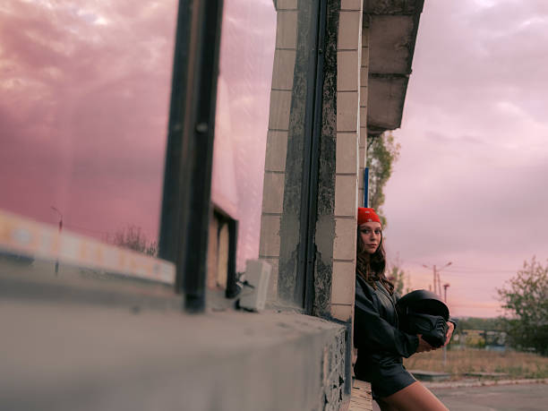 Young, stylish biker woman in leather jacket, shorts and bandana leans against an abandoned building, holding her helmet. Striking reflection of sunset sky in windows.