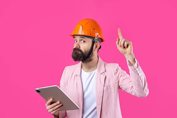Portrait of young construction engineer wear orange hard hat, in a pink jacket standing on red studio background. A man with a tablet in his hands. stock photo