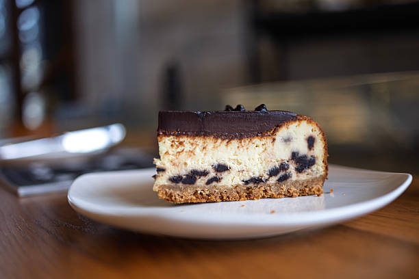 Close-up of a cheesecake slice with chocolate topping and cookie crust, served on a white plate in a cafe.