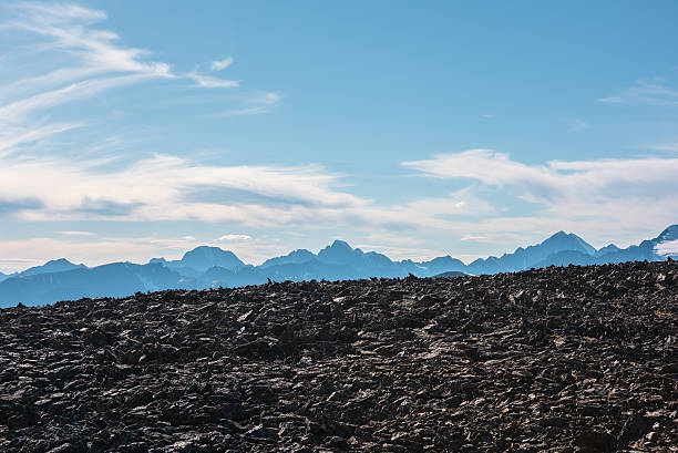 Top view from sunlit stony plain to large rocky mountains silhouettes with pointy peaks in bright sun under clouds in blue sky. Big silhouette of mountain range with peaked tops far away in sunny day.