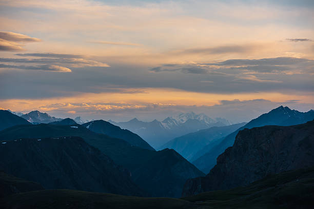 Deep gorge between layered silhouettes of sharp rocky ridge spurs under huge snowy mountain peak in golden cloudy sky. Sheer crags of mount valley under giant ice top under gold sunset color clouds.
