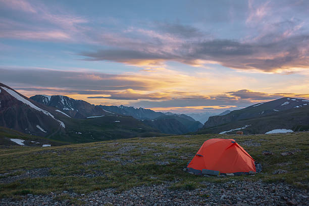 istock Orange tent on grassy stony pass with view to mountain range silhouette far away under clouds in vivid sunset color sky. Scenic alpine landscape with red tent in mountains under colorful cloudy sky. 2231654895