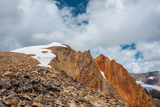 istock Snow-capped mountain ridge with cornice and red sharp rocks on top in sunlight. Snow cap and shiny pointy peak of gold color. Snow dome and colorful rocky peaked top. Sunny cloudy high mountains view. 2231655841