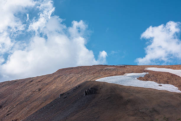 istock Rock and snowfield on mountainside in changeable weather. Sunlit stony mountain range with snow under clouds in blue sky. Sunlight and shadows on stone hill in sunny day. Rocky hillside in bright sun. 2231656517