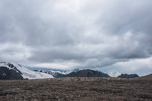 Dramatic misty top view from stone pass to big glacier tongue among sharp rocks and large snow-capped pinnacle in rainy low clouds. Dark atmospheric mountain silhouettes in rain in gray cloudy sky.
