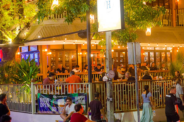 Manhy customers inside and outside of bar bistro Hemingway in Bangkok street Sukhumvit Road Soi 11 at night. View over street. People are sitting in front yard or inside. In foreground people and tourists are walking along sidewalk. At corner at fence is a banner wwith happy beer hour. oi 11 is popular nightlife place for tourists and western people in Bangkok downtown with many bars and soime restaurants