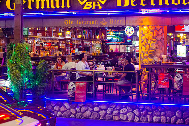 Tourists are sitting on  a beer break in bar. Bar is called German Beerhouse. Scene is in street Sukhumvoit Road Soi 11. Soi 11 is popular nightlife place for tourists and western people in Bangkok downtown with many bars and some restaurants. Main focus is on man holding a bottle of beer. On his t-shirt is the text \