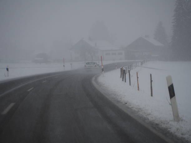Snowy road with a car driving down it. The car is white and is in the middle of the road. The road is wet and slippery