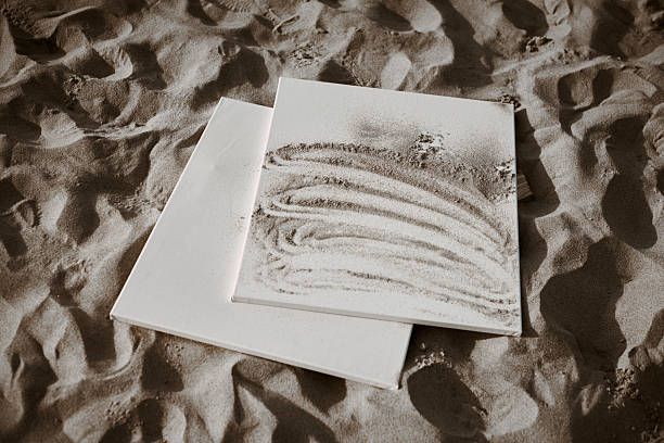 Two white canvases lie on sandy surface, one featuring intricate sand patterns. Scene evokes sense of tranquility and creativity, capturing essence of art in nature