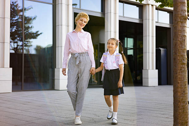 A mother and her elementary student daughter walk together in a schoolyard, holding hands and smiling.