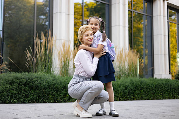 A mom embraces her daughter in front of a school building. The young girl wears a backpack, ready for the school day.