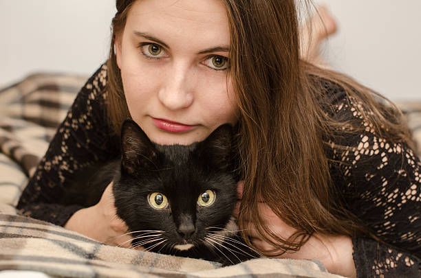 Young woman with long brown hair lies on plaid blanket beside black cat with white patch. Both share calm moment, eyes directed at camera, soft indoor light enhancing scene