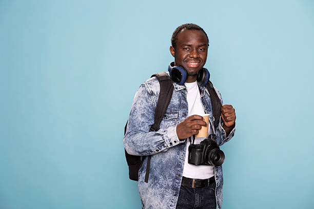 Black man in relaxed pose, gripping his backpack, holding a coffee cup, with wireless headphones resting around his neck. Portrait of african american photographer standing against isolated background.