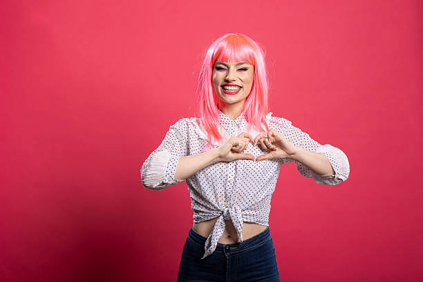 Happy pink haired woman in casual clothing forms a heart shape with her hands, expressing love and joy. Beautiful young lady standing isolated on pink background with romantic smile.