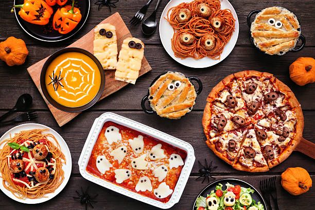 Spooky Halloween dinner party table scene over a dark wood background. Above view. Skull pizza, salad, pasta, ghost lasagna, mummy pot pie, pumpkin soup, jack o lantern stuffed peppers.