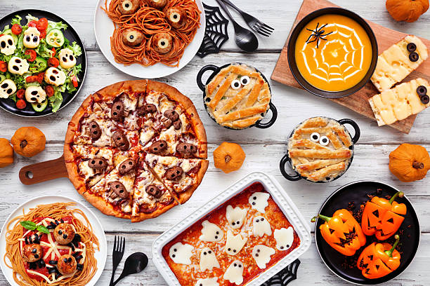 Spooky Halloween dinner party table scene over a white wood background. Top view. Skull pizza, salad, pasta, ghost lasagna, mummy pot pie, pumpkin soup, jack o lantern stuffed peppers.