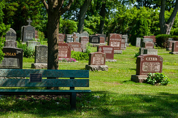 Burlington, Ontario, Canada - June 12, 2024: Serene cemetery landscape featuring gravestones surrounded by lush greenery, complemented by a memorial bench under a tree, emphasizing respect, remembrance, and peace.