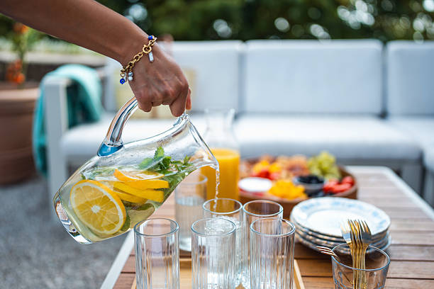 A person is pouring water from a glass pitcher filled with lemon and mint into glasses on a wooden table in a beautiful backyard setting. The table is featuring a spread of fruits and a sofa in the background.