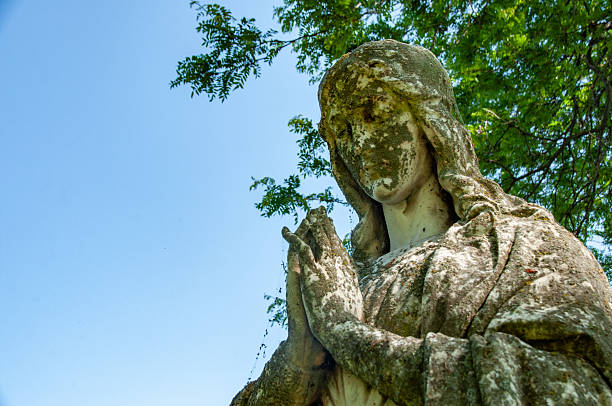 A weathered stone statue of a praying figure, surrounded by natural green foliage, viewed from below, emphasizing its serene presence and ancient beauty against the bright sky.