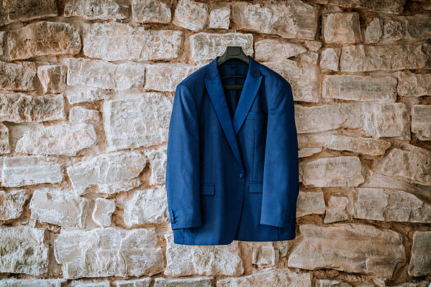 istock A stylish blue men's suit jacket hangs on a hanger against a rustic stone wall, ready for a formal event or wedding day. 2231698111