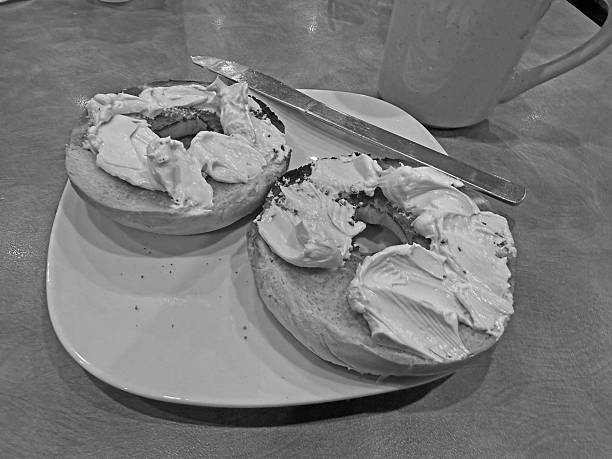A toasted, sliced, plain bagel on a white plate.   Cream cheese spread on both slices of the bagel.    Stainless steel knife on the edge of the square plate with rounded edges.   Coffee cup on the upper right side of photograph.   Black and white photograph.