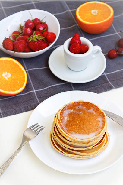 Bright breakfast spread: golden pancakes with syrup beside halved oranges, cherries, and strawberries, set on a white table with a gray napkin, cup, fork, and knife