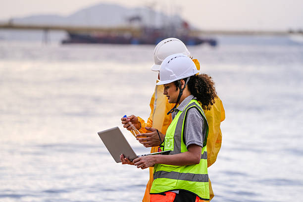 Two field scientists in PPE gear analyzing seawater samples using test tubes and a laptop. Environmental monitoring and pollution control on the coastline are depicted.