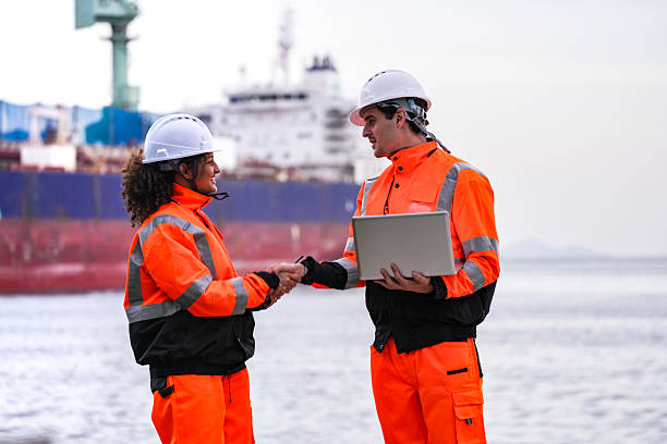 istock Two port engineers in high-visibility orange uniforms and hard hats communicate while inspecting dockside logistics using a laptop and walkie-talkie, symbolizing teamwork and safety. 2231739755