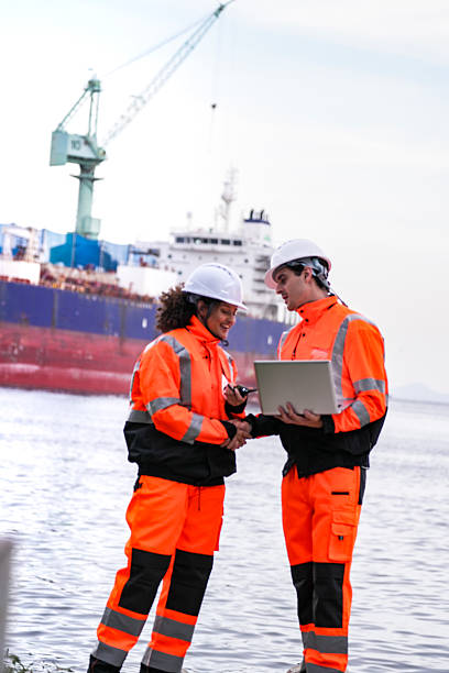 istock Two port engineers in high-visibility orange uniforms and hard hats communicate while inspecting dockside logistics using a laptop and walkie-talkie, symbolizing teamwork and safety. 2231740047